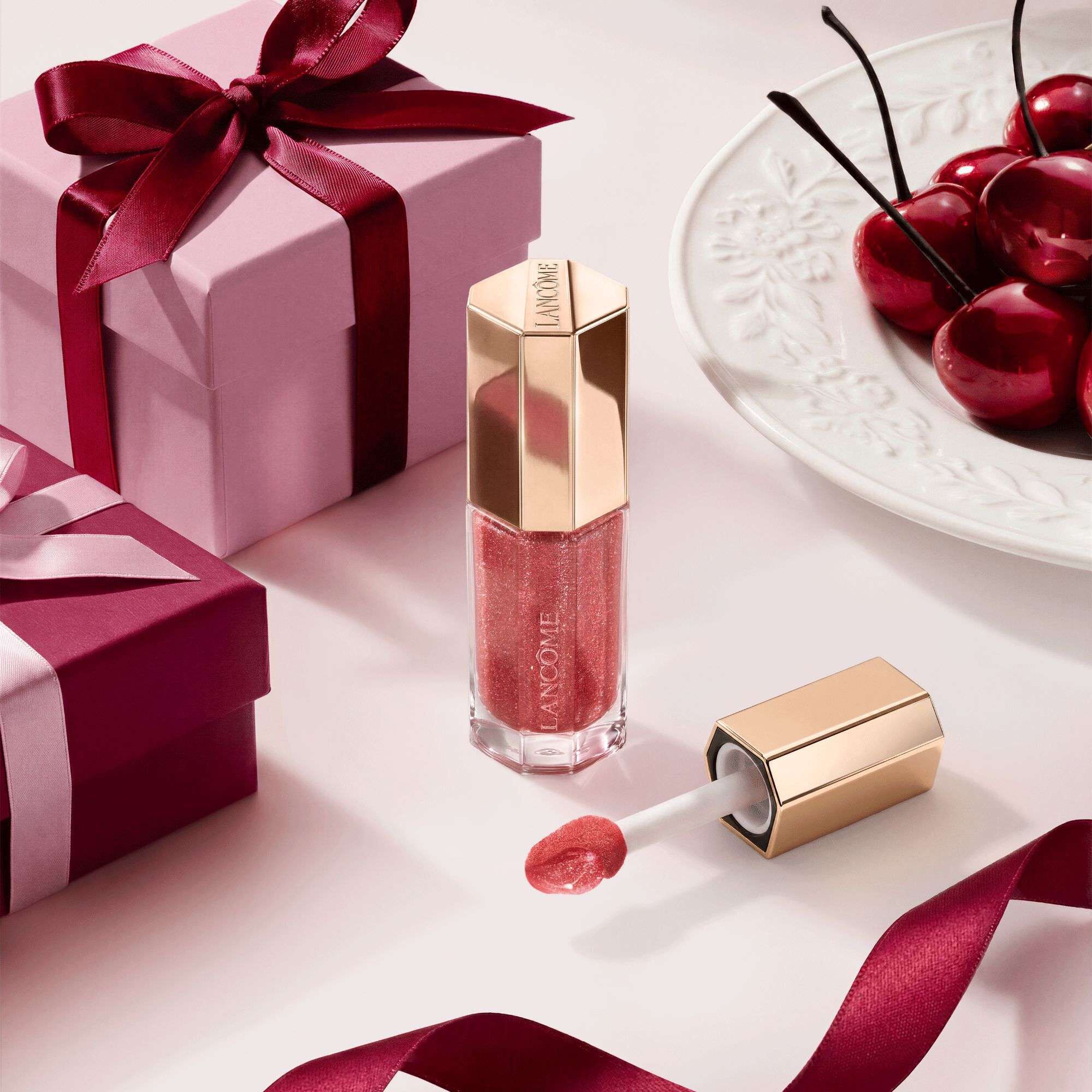 Lancome berry-toned lip gloss in a luxury hexagonal tube, styled with gift boxes and cherries.	