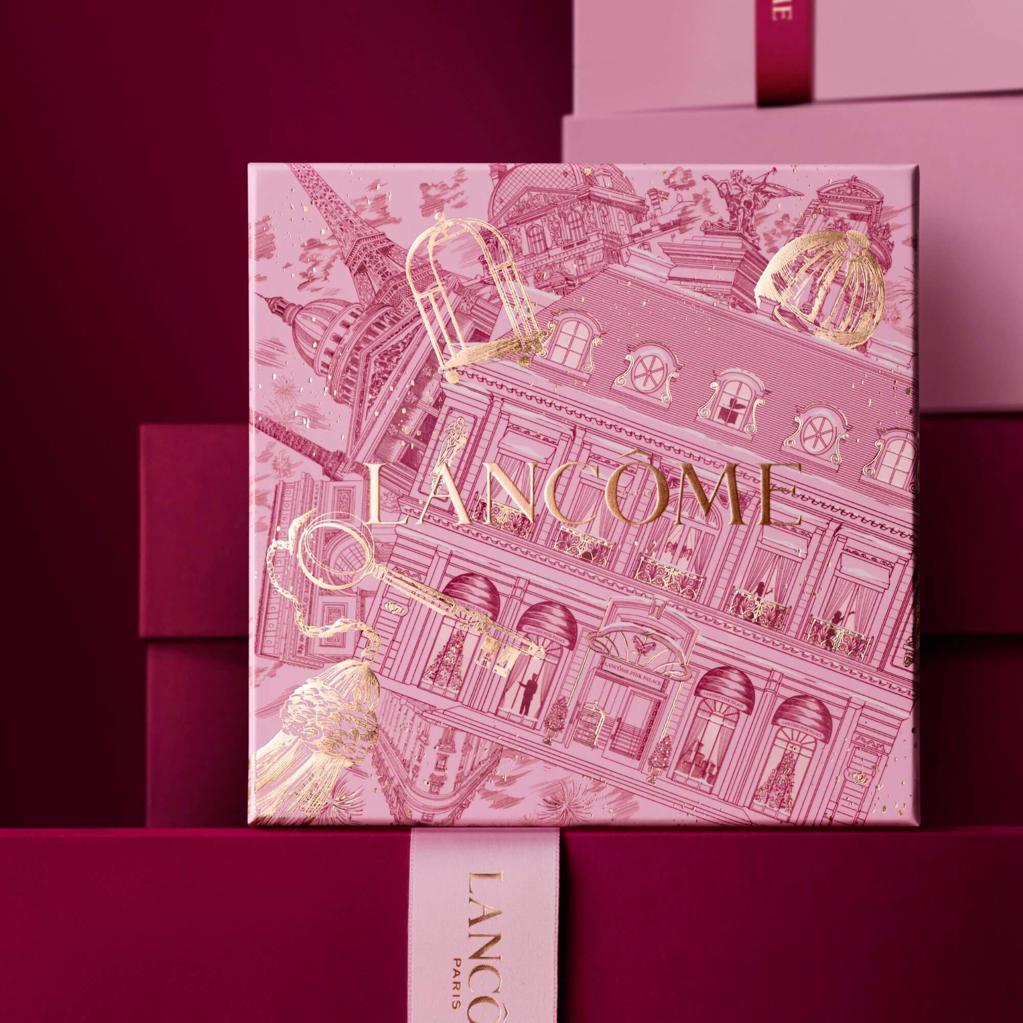 Image of pink Lancome holiday gift box standing in front of a pile of pink and dark red gift boxes, set against a dark red background. 