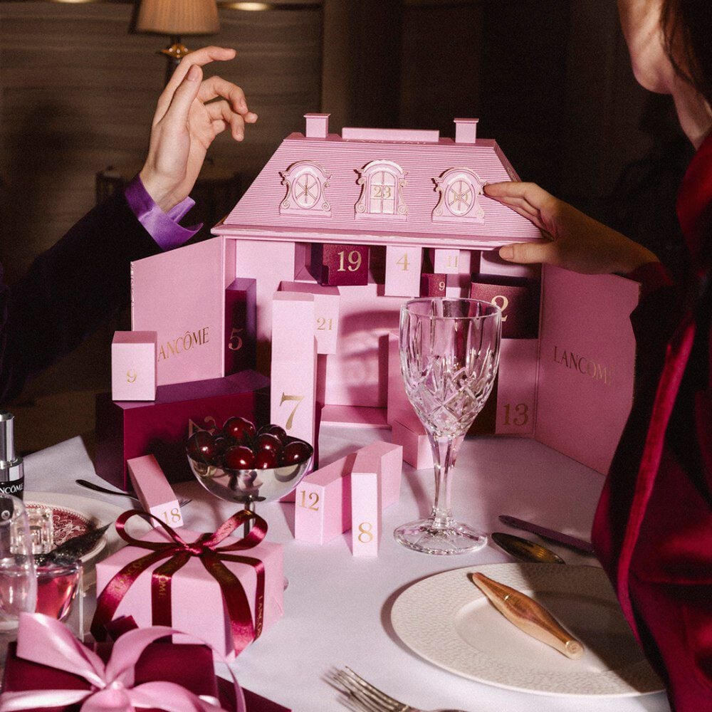 Hands interacting with a pink Lancome advent calendar on a festive dining table with tableware and cherries.